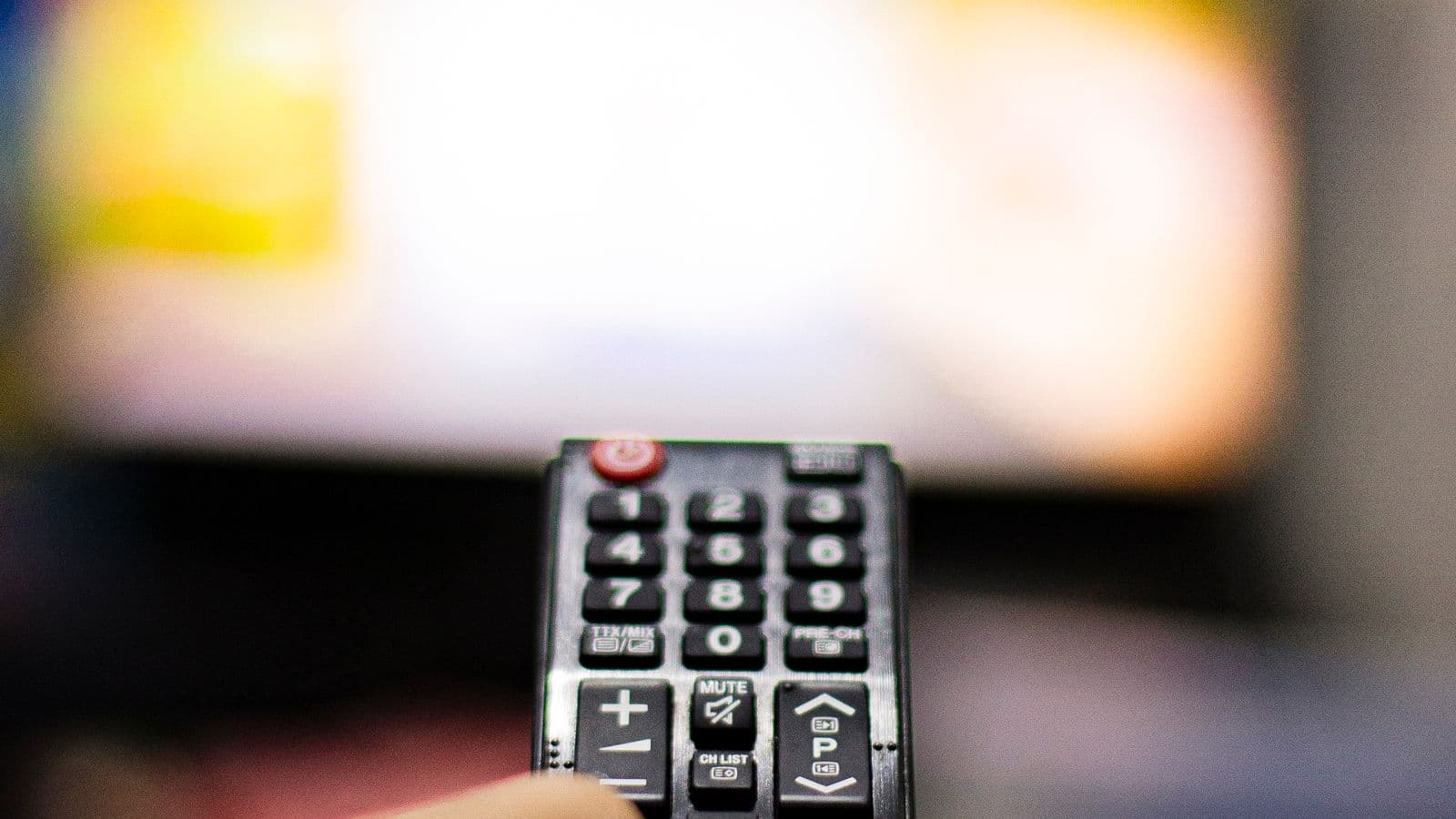 Pilot do telewizora z klawiszami numerycznymi skierowany w stronę rozmytego, jasnego ekranu telewizora
