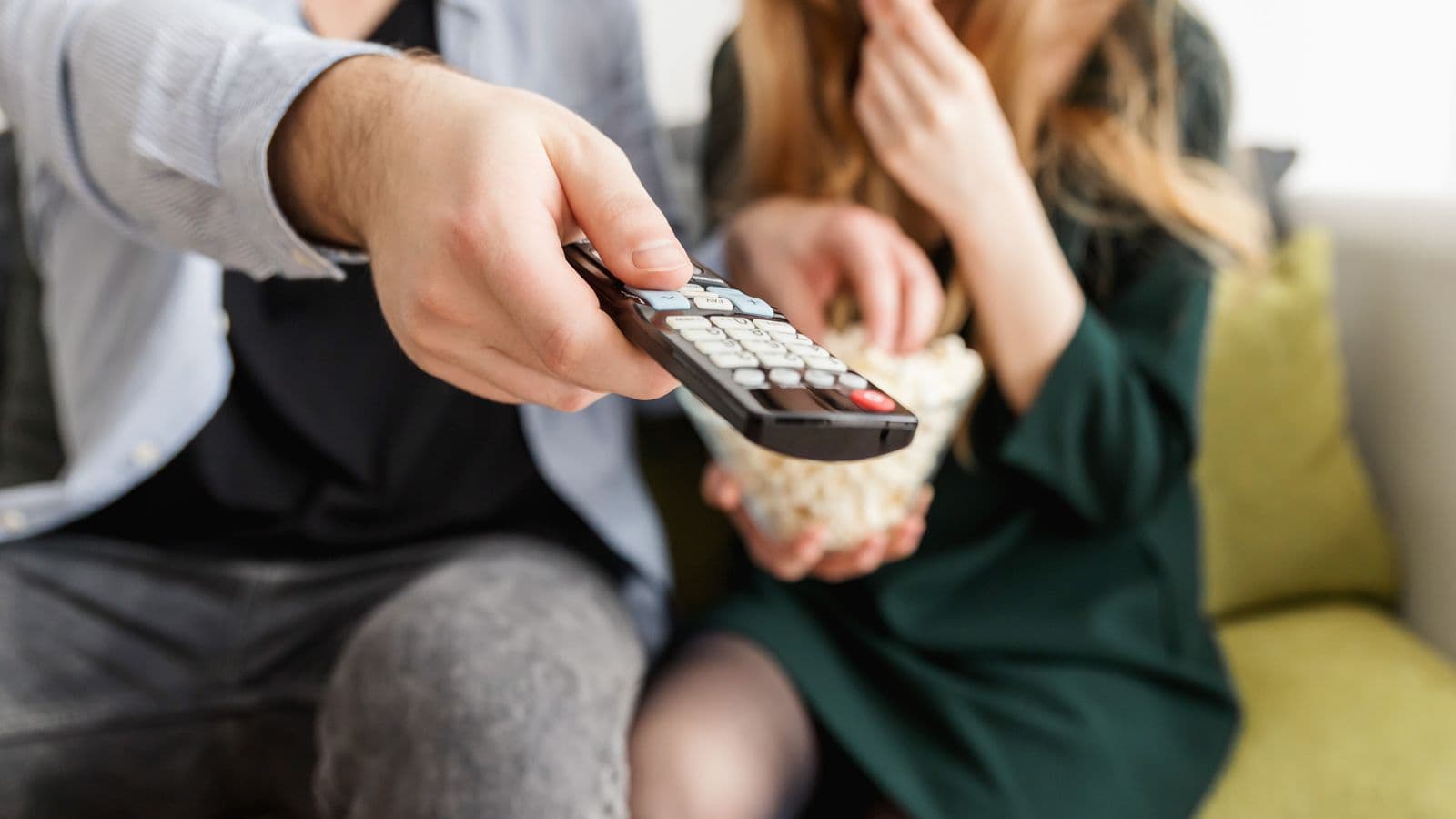 Dłoń kierująca pilota do telewizora, w tle dwie osoby na kanapie z miską popcornu